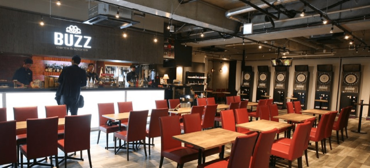 BUZZ darts&sports 船橋店の店内全体・赤い椅子のダイニングテーブル席とバーカウンター、ダーツマシン