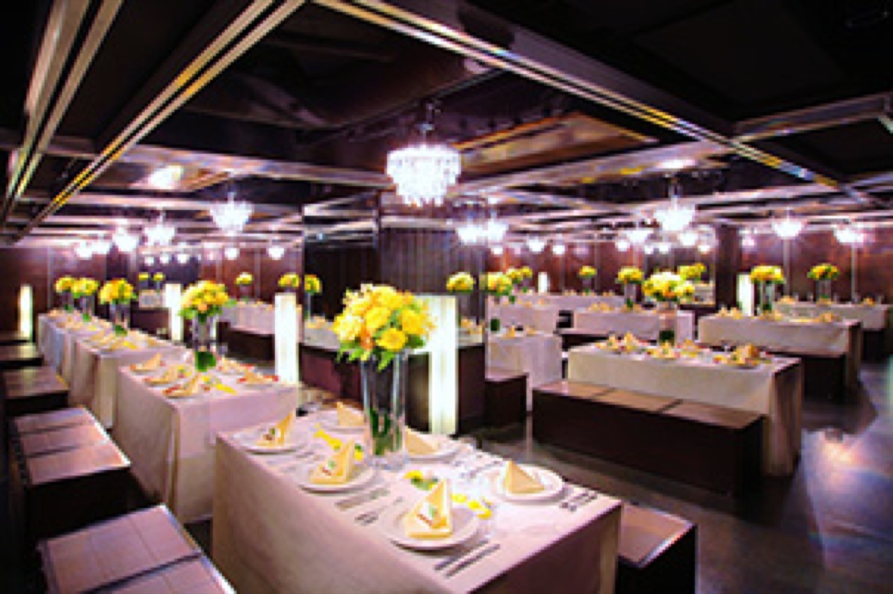 PARTY For ETERNITY 銀座店の宴会場内観。白いテーブルクロスと黄色い花を飾ったテーブルセッティングが整った、シャンデリア照明の明るい貸切パーティールーム（96名様用・披露宴正餐レイアウト）。