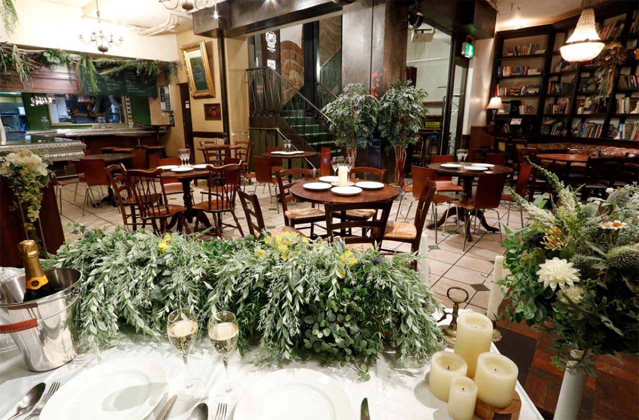 Green Lounge 渋谷の会場内観。観葉植物のグリーンで装飾されたダイニングフロアに木製の丸テーブルと椅子が並び、奥にバーカウンターと本棚、シャンデリアが配されたラウンジ空間